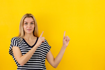 Amazing blonde girl in striped t-shirt points fingers up, smiling and looking at camera on yellow background. Copy space for your promo