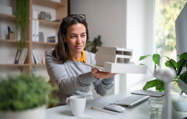 Architect with model of a house sitting at the desk indoors in office. - obrazy, fototapety, plakaty