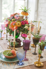 Festive table in boho chic style. Decorated with fresh flowers of peonies, lilies, succulents. Cutlery and plates on a wooden table, candles in bronze vintage coasters and colored glasses for wine.