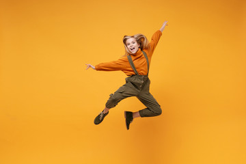 Fototapeta na wymiar Cheerful little kid girl 12-13 years old in turtleneck, jumpsuit isolated on orange yellow wall background in studio. Childhood lifestyle concept. Mock up copy space. Jumping spreading hands and legs.