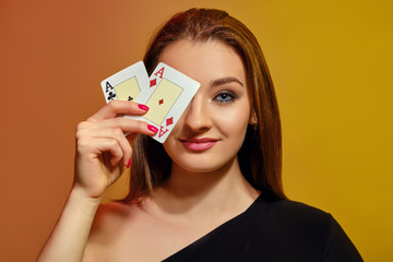 Blonde girl in black dress has covered her eye with two playing cards, smiling, posing on colorful background. Poker, casino. Close-up.