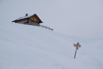 Lonely small wooden house and tourist signpost in snowy mountains in foggy winter day, when all around is white. Around Alpe di Nemes refuge in Sexten Dolomites, South Tyrol, Italy