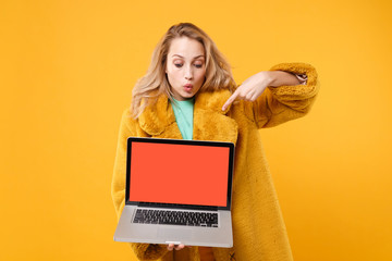 Naklejka premium Amazed young blonde woman in yellow fur coat isolated on orange background in studio. People lifestyle concept. Mock up copy space. Pointing index finger on laptop pc computer with blank empty screen.