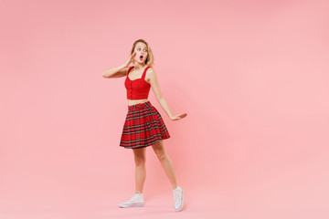 Shocked young blonde woman girl in red sexy clothes isolated on pastel pink background. People lifestyle concept. Mock up copy space. Whispering secret behind her hand, sharing news, looking aside.