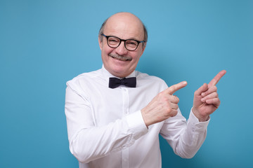 Good looking senior man with satisfied expression, indicates with fore fingers aside on copyspace,...