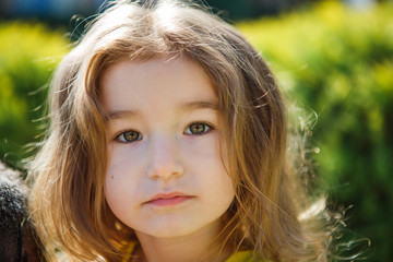A 3-year-old girl with long, lush blond hair looks into the frame. Small blonde close-up with a serious face in the background light