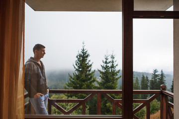 A young man in the morning looks from the hotel balcony with a view of the mountains, the fog and...