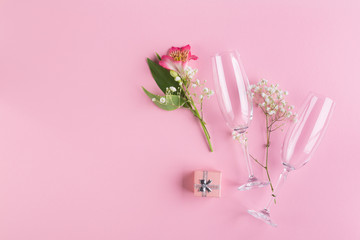 A bouquet of pink and white flowers, a small gift box and two glasses for sparkling wine on a pink paper background.