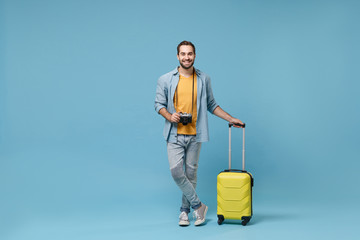 Smiling traveler tourist man in yellow casual clothes with photo camera, suitcase isolated on blue background. Male passenger traveling abroad on weekends. Air flight journey concept. Looking camera.