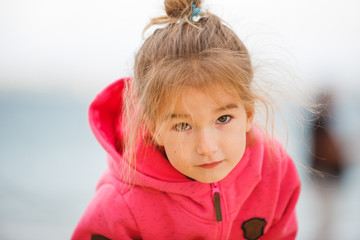A 4-year-old girl with blonde hair in a bun with her hair flying in the wind in a pink hoodie.  Girl close-up, with a blurred background, playing near the road