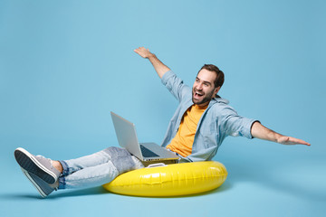 Funny traveler tourist man in yellow clothes isolated on blue background. Passenger traveling abroad on weekend. Air flight journey concept. Sitting in inflatable ring work on laptop, spreading hands.