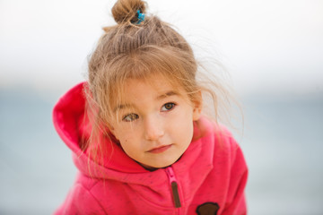 A 4-year-old girl with blonde hair in a bun with her hair flying in the wind in a pink hoodie.  Girl close-up, with a blurred background, playing near the road