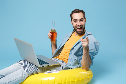 Traveler Tourist Man In Yellow Clothes Isolated On Blue Background. Passenger Traveling Abroad On Weekend. Air Flight Journey Concept. Sit In Inflatable Ring Work On Laptop Hold Beer Point On Camera.