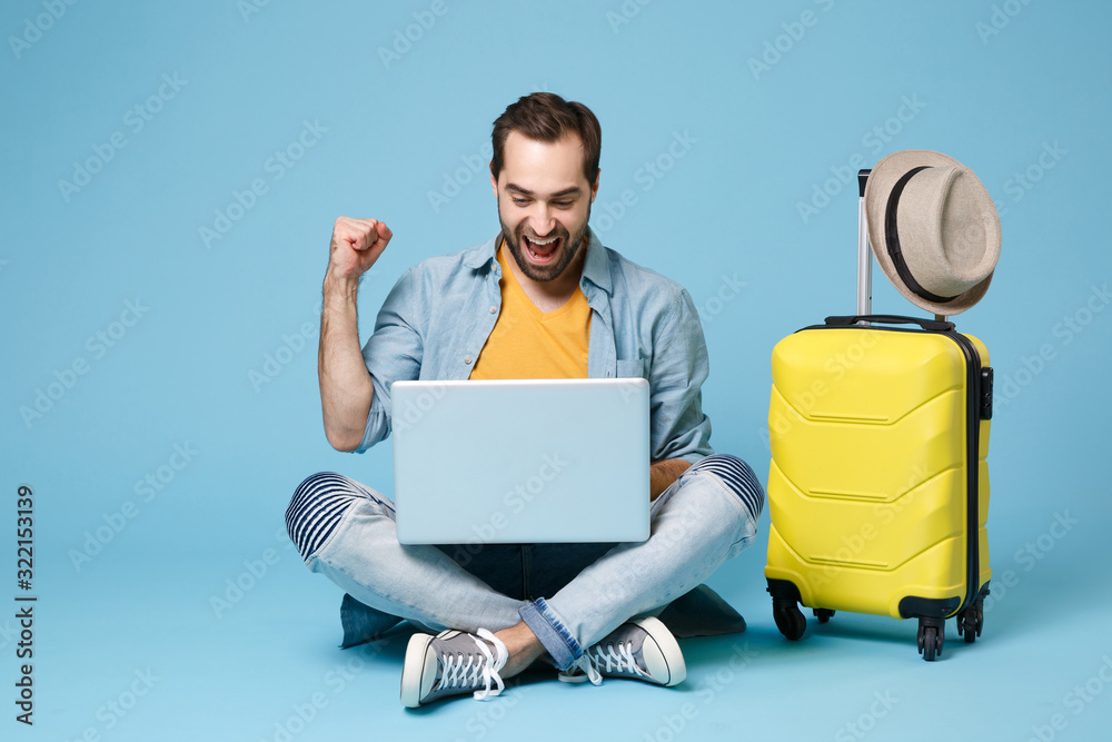 Wall mural Joyful traveler tourist man in yellow clothes isolated on blue background. Passenger traveling abroad on weekend. Air flight journey concept. Sit near suitcase, work on laptop, doing winner gesture.