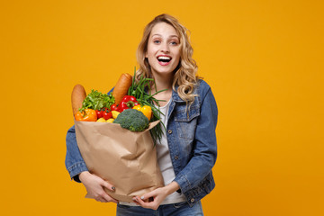 Cheerful young woman in denim clothes posing isolated on yellow orange background. Delivery service from shop or restaurant concept. Hold brown craft paper bag for takeaway mock up with food products.