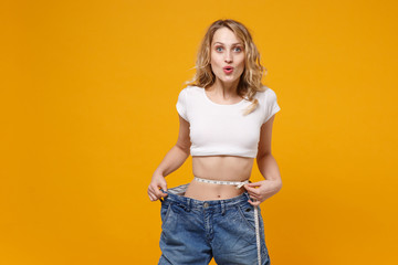 Amazed young woman in white t-shirt isolated on yellow orange background. Proper nutrition losing weight healthy lifestyle dieting concept. Wearing old big jeans measuring waist with measure tape.