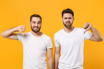 Confused displeased young men guys friends in white blank empty t-shirts posing isolated on yellow orange background studio portrait. People lifestyle concept. Mock up copy space. Showing thumbs down.