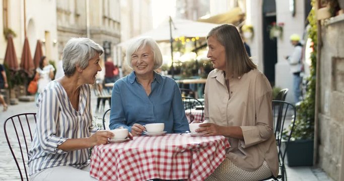 Three Caucasian senior women friendly chatting and drinking coffee at table in cafe terrace in summer. Female friends talking and laughing outdoor while gossiping and sipping tea.