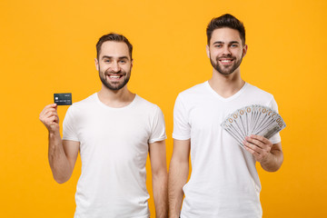 Smiling young men guys friends in white blank t-shirts isolated on yellow orange background. People lifestyle concept. Mock up copy space. Hold fan of cash money in dollar banknotes, credit bank card.