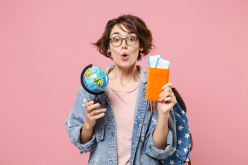 Amazed young woman student in denim clothes eyeglasses, backpack posing isolated on pastel pink background. Education in high school university college concept. Holding passport tickets world globe.