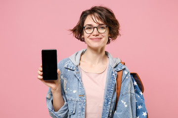 Smiling young woman student in denim clothes, eyeglasses, backpack isolated on pastel pink background. Education in high school university college concept. Hold mobile phone with blank empty screen.