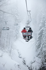 Two snowboarders sitting on the ski chair lift. Ski lift in the forest. Extreme snow conditions,...