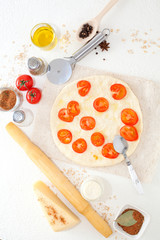 Ingredients for pizza, on a white table a knife for slicing, a rolling pin for dough, spices and black pepper peas, next to a half-assembled pizza decorated tomatoes. Concept making pizza.