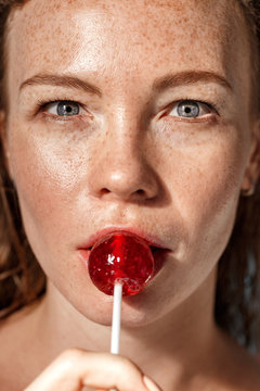Summer Freestyle. Young woman with freckles standing isolated on blue licking lollipop close-up looking camera curious