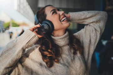 Young happy woman listening to music in headphones outdoors, positive hipster girl in cozy knitted sweater enjoying sunny day, Good vibes  Lifestyle concept
