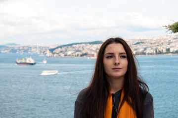 Fototapeta na wymiar Attractive brunette with an uneasy look looking into the distance. Bosphorus canal in Istanbul, turkey behind her , blurred ships and istanbul peninsula