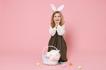 Obraz na płótnie Canvas Little pretty blonde kid girl 11-12 years old in light spring dress hold fluffy white bunny rabbit, wicker basket with eggs isolated on pastel pink background. Childhood lifestyle Happy Easter concept