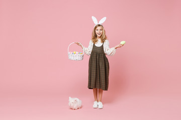 Fototapeta premium Little pretty blonde kid girl 11-12 years old in light spring dress hold fluffy white bunny rabbit, wicker basket with eggs isolated on pastel pink background. Childhood lifestyle Happy Easter concept