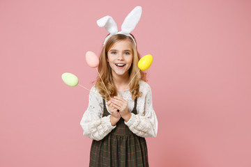 Obraz na płótnie Canvas Little pretty blonde kid girl 11-12 years old in spring dress, bunny rabbit ears hold in hand carries dyed eggs on sticks have fun celebrate isolated on pastel pink background. Happy Easter concept.