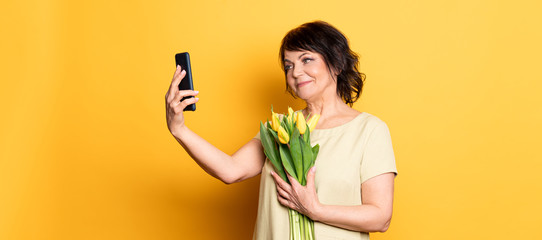 Beautiful old woman with professional smokey make-up and hairstyle holding tulip flowers on the pink background. Taking video call by the smartphone .Concept spring