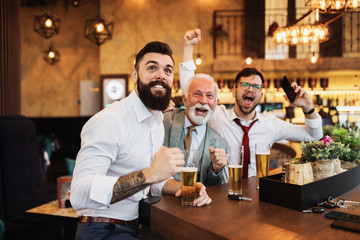  Three businessmen cheering while watching football match in a cafe bar.