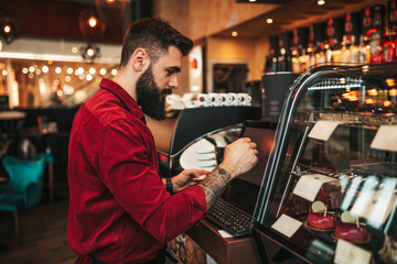 Young handsome bearded barista in red shirt working  in modern cafe bar.