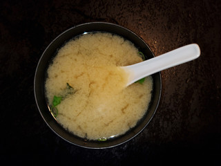 Top down view of bowl of Miso soup with spoon garnished by green onions on dark textured background
