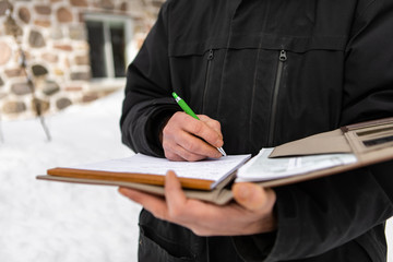 selective focus and close up view of man's hands as he holds a notebook and writes notes, home inspector wearing winter clothes outdoor in snow