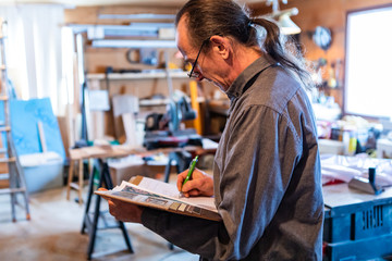 Fototapeta na wymiar middle aged man with long hair wearing glasses in the garage with tools in the background. home inspector taking professional notes on his notebook
