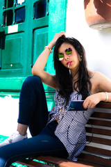 Colombian woman wears sunglasses and listens to music from her cell phone with headphones sitting on a wooden chair eating a red candy