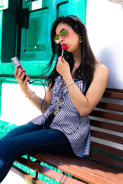 Colombian woman wears sunglasses and listens to music from her cell phone with headphones sitting on a wooden chair eating a red candy