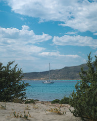 Sailboat seen from between green bushes in Elafonisos, Greece on a beautiful day with blue sky and clouds.