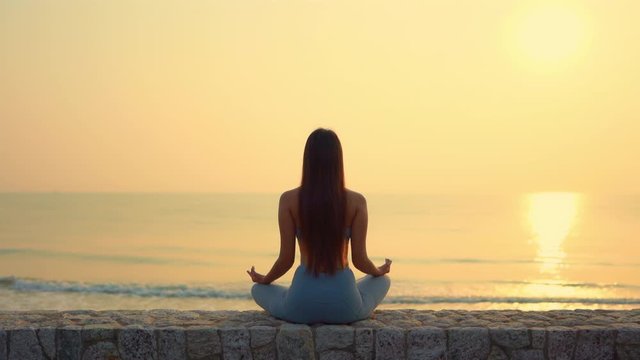 Slim woman practicing Yoga by meditating in Zen position near the ocean during a golden sunset.