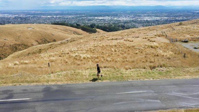 Profile tracking shot of man running on grass by road on hill.