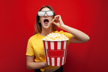 beautiful young emotional girl in 3D glasses eats popcorn, watches a movie and screams on a red color background, surprised viewer