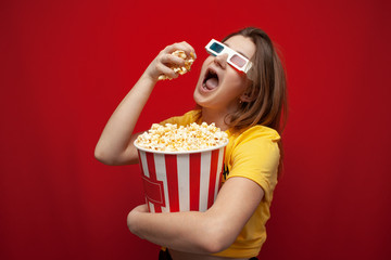 beautiful young girl in 3D glasses eating popcorn and watching a movie on a red color background