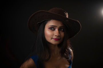 Fashion portrait of young and attractive Indian Bengali brunette girl with blue western dress with cowboy hat under spotlight in front of black studio background.Indian fashion portrait and lifestyle.