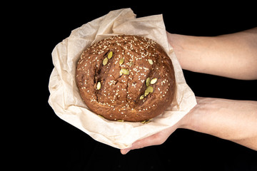 Man's hands hold tasty fresh loaf of dark bread with sesame seeds on black background