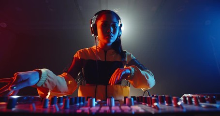 Cool asian female disc jockey standing at mixer cotroller, creating a music list in a nightclub, lit by red and blue lights - nightlife concept 4k footage