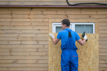 a worker in a blue uniform, blocks the window of the house with a protective shield made of wood, from thieves, when moving to another address, there is a place on the left for the inscription